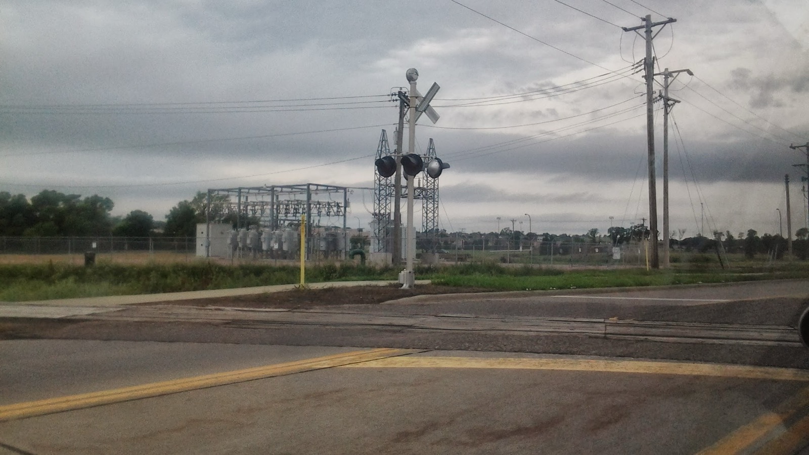 Railroad crossing signals and a crossbuck beside tracks and a road in Yankton, South Dakota, with an electrical substation behind them.